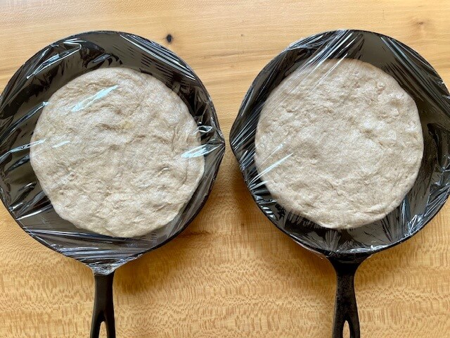 two cast iron pans with fresh milled flour pizza crust in them and covered with plastic wrap