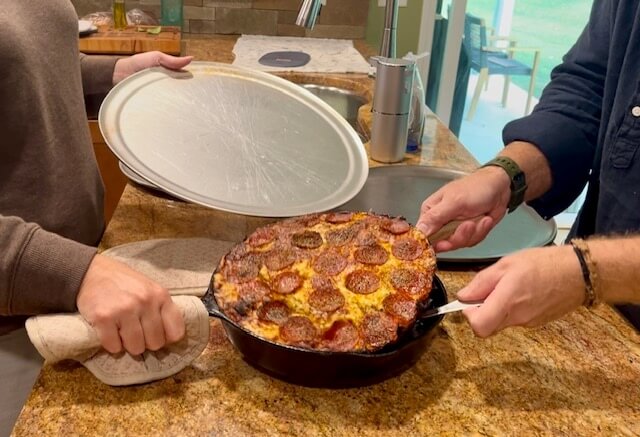 taking a pepperoni fresh milled flour pizza out of a cast iron pan with fish turners to put on a pizza pan