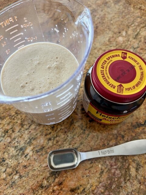 Bubbly yeast in liquid in a measuring cup next to the bottle of yeast and a measuring teaspoon for fresh milled english muffins