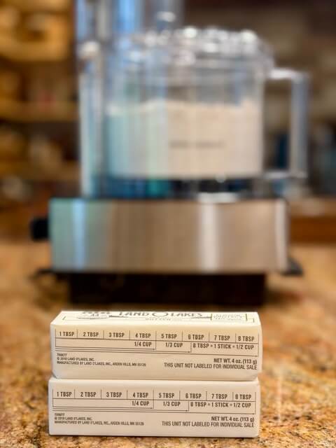Two sticks of butter on the counter with the food processor in the background for fresh milled flour pie crust recipe