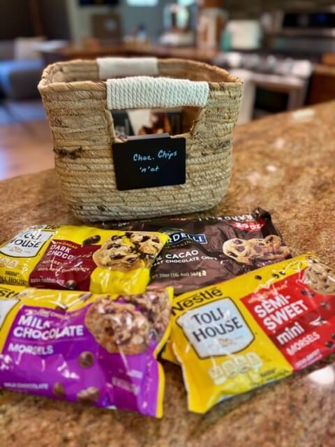 Bags of different kinds of chocolate chips on counter in front of a basket for putting into fresh milled flour brownies