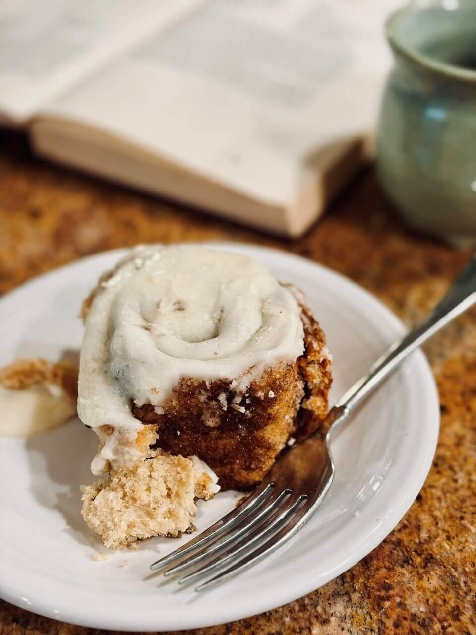 Fresh Milled Cinnamon Roll on a white plate next to a book and cup of coffee