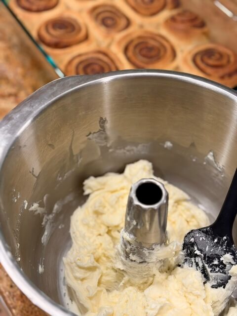 A bowl of frosting in front of a pan of fresh milled cinnamon rolls